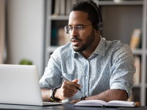 Man wearing headphones and glasses works at a laptop, taking notes in a notebook with a pen nearby in a home office.