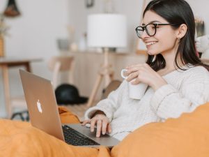 Smiling woman wearing glasses uses a laptop on a couch, holding a mug in a cozy living room.