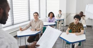 Classroom scene with a teacher at the front and five students seated at individual desks, each with papers.