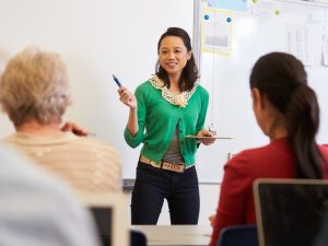 Instructor in a green cardigan stands at the front of a classroom, gesturing with a marker while holding a clipboard near the whiteboard.