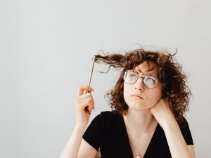 Woman with curly hair and round glasses holds a pencil to her hair, looking upward in thought.