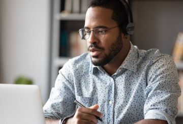 Man wearing headphones and glasses works on a laptop, holding a pen at a desk in a home office