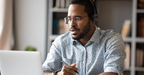 Man wearing headphones and glasses works on a laptop, holding a pen at a desk in a home office