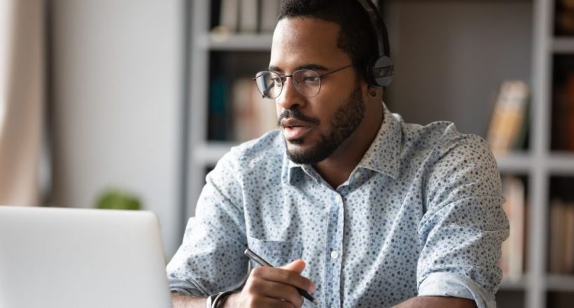 Man wearing headphones and glasses works on a laptop, holding a pen at a desk in a home office