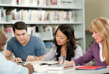 Group of four adults studying together at a library table with books and notebooks open in front of them, collaboratively reading.