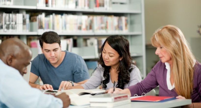 Group of four adults studying together at a library table with books and notebooks open in front of them, collaboratively reading.