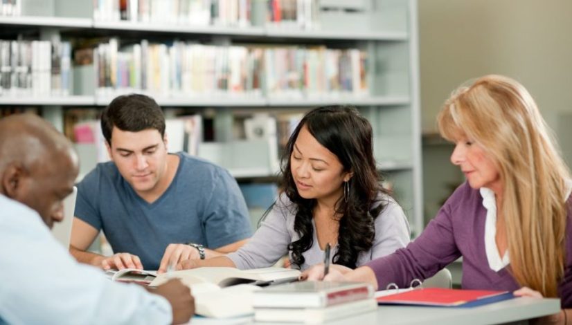 Group of four adults studying together at a library table with books and notebooks open in front of them, collaboratively reading.