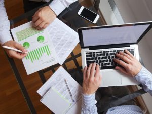 Two professionals collaborating over charts and a laptop on a glass table, with a smartphone nearby.
