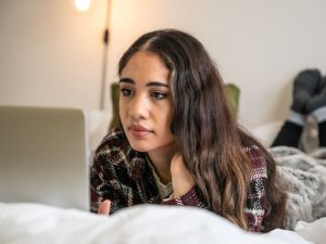 Young woman lying on a bed, propped on her elbow, looking at a laptop screen at home in a cozy bedroom.