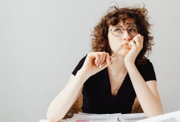 Woman with curly hair and round glasses sits at a desk with scattered papers, holding a pencil to her lips as she thinks.