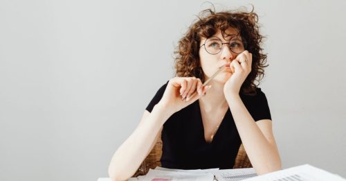 Woman with curly hair and round glasses sits at a desk with scattered papers, holding a pencil to her lips as she thinks.