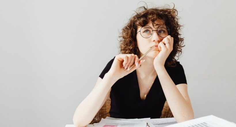 Woman with curly hair and round glasses sits at a desk with scattered papers, holding a pencil to her lips as she thinks.