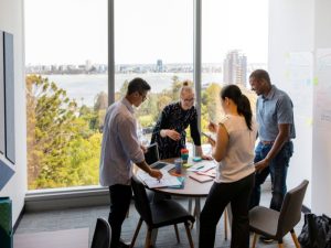 Four colleagues stand around a round table in a bright office, reviewing papers as they brainstorm, with a city and water view through a large window behind them.