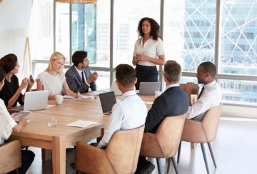 Diverse group of professionals in a modern office listening to a woman presenting at a conference table.