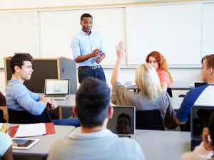 Instructor standing at front of classroom as students with laptops listen; a student raises a hand.