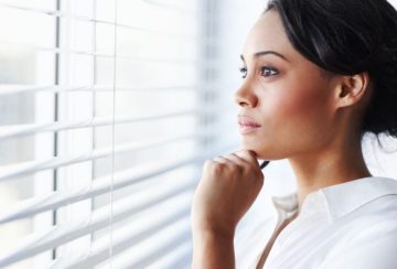 Profile of a woman in a white blouse gazing thoughtfully out of window blinds, hand resting on chin