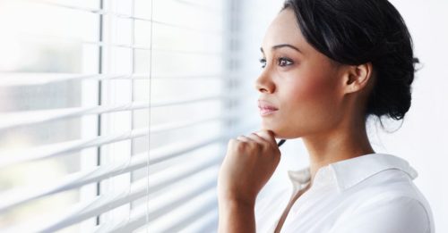 Profile of a woman in a white blouse gazing thoughtfully out of window blinds, hand resting on chin